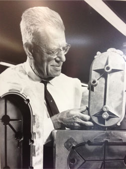 Carpenter Die Casting An older man in glasses and a tie examines a mechanical component, surrounded by industrial equipment at Carpenter Die Casting.