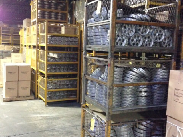 Carpenter Die Casting Industrial storage area with metal shelving holding various metal parts and wire baskets, surrounded by cardboard boxes and crates on a concrete floor.