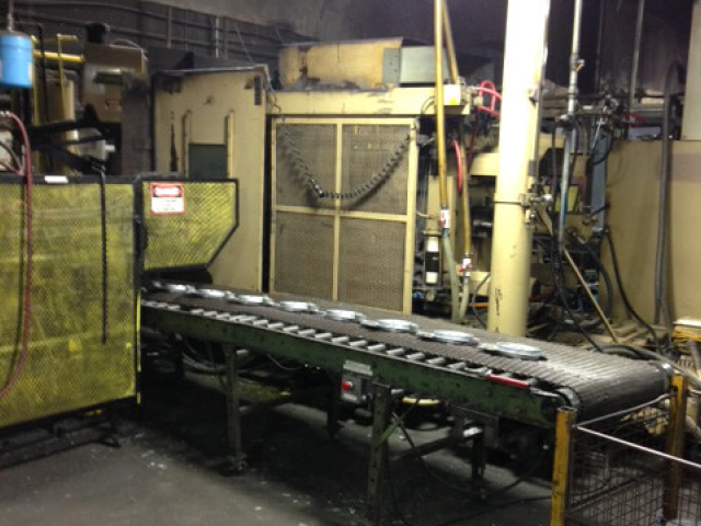 Carpenter Die Casting A factory conveyor belt transports several round metal objects through an industrial processing area with machinery and pipes in the background.