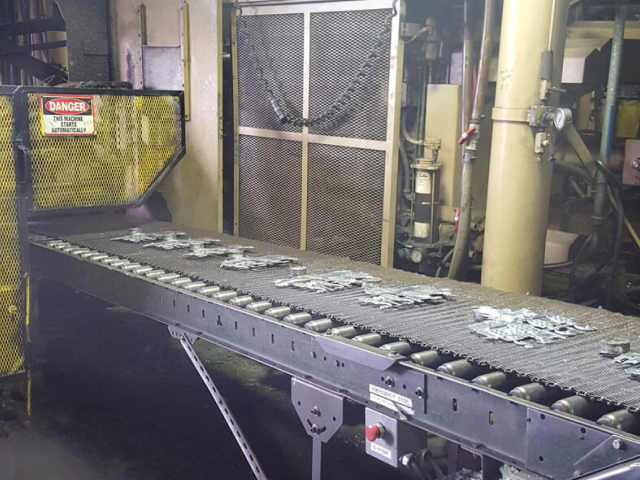 Carpenter Die Casting Metal parts are moving along a conveyor belt in an industrial factory setting with a "Danger" sign visible on a machine guard.