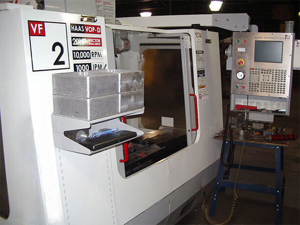 Carpenter Die Casting A Haas VF-2 vertical machining center with its door open, control panel visible on the right, and metal parts on a shelf beside the machine.