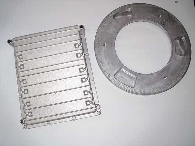 Carpenter Die Casting Two machined metal parts, one rectangular with ridges and mounting holes, and one circular with embossed text and cutouts, are displayed on a white surface.