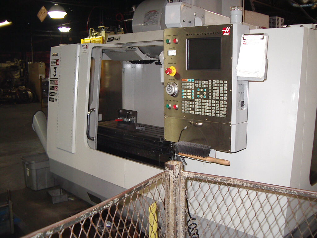 Carpenter Die Casting A CNC milling machine in an industrial workshop, with its control panel and open working area visible behind a chain-link barrier.