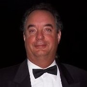 Carpenter Die Casting A man wearing a black tuxedo and bow tie stands against a dark background, facing the camera and smiling slightly.