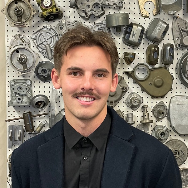 Carpenter Die Casting A young man with short brown hair and a mustache stands in front of a pegboard wall covered with various metal mechanical parts. He is wearing a dark coat and a black shirt.