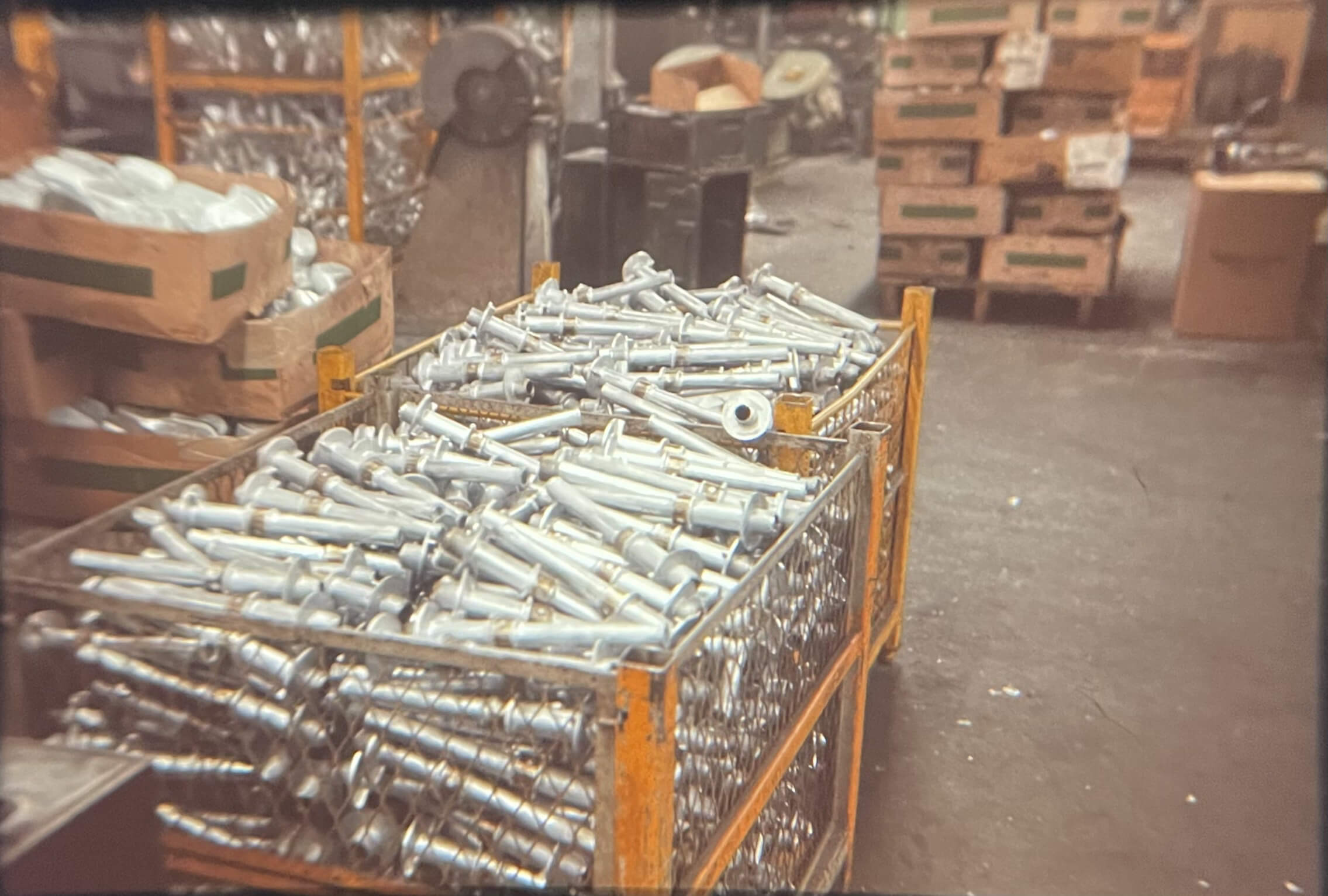 Carpenter Die Casting Large yellow metal crate filled with silver metal rods or bolts in a warehouse with boxes and industrial materials in the background.