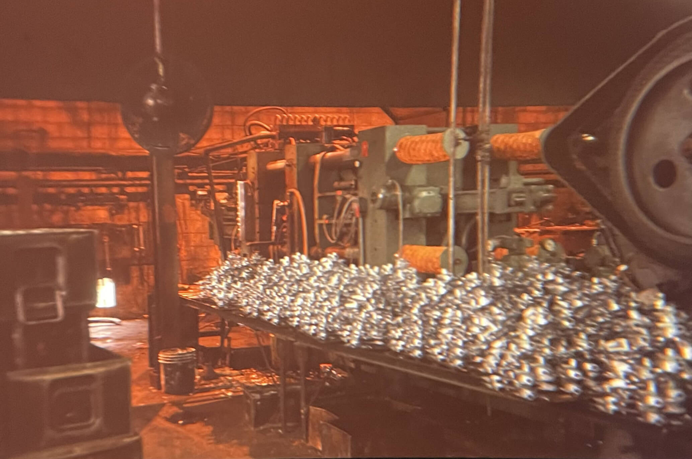 Carpenter Die Casting A pile of shiny metal parts sits on a conveyor belt inside an industrial factory with heavy machinery and dim lighting.