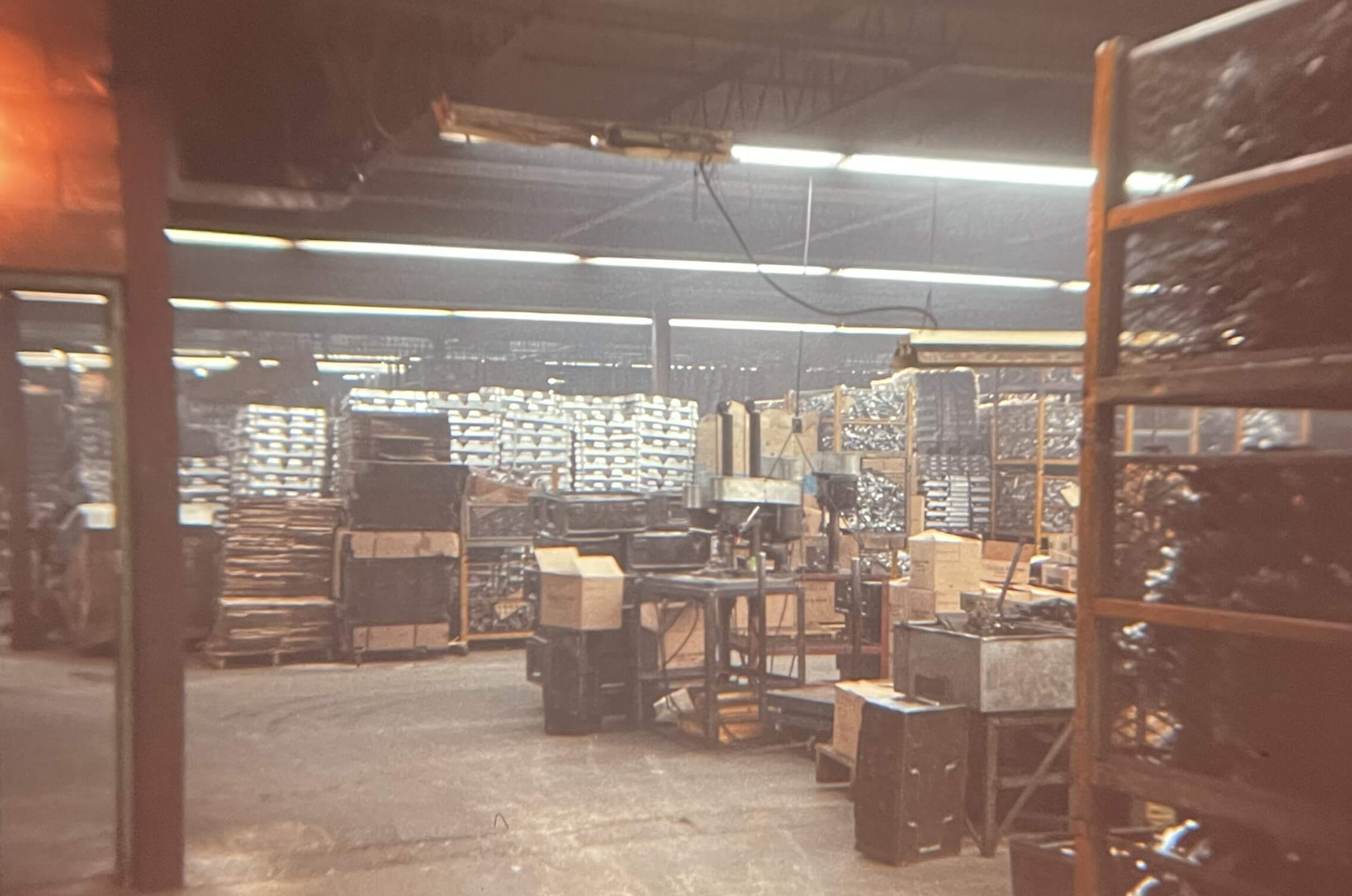 Carpenter Die Casting A dimly lit warehouse interior with shelves stacked with boxes and metal parts, industrial equipment, and scattered crates and tools.