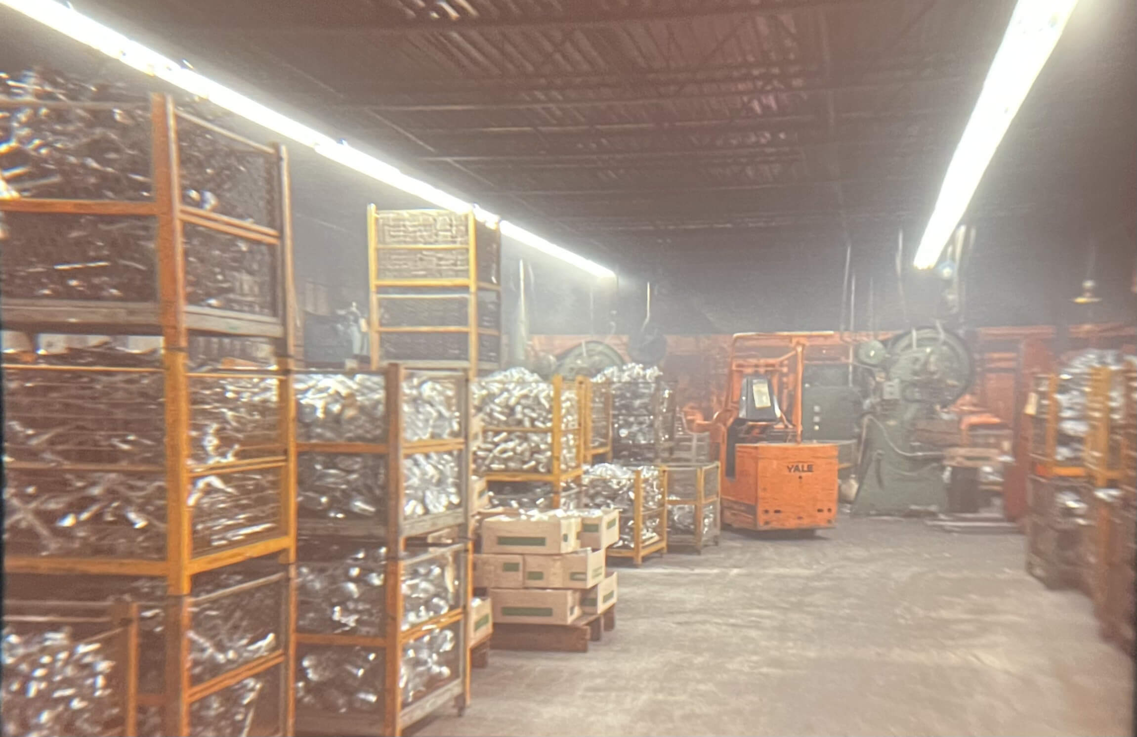 Carpenter Die Casting Stacks of metal parts in wire bins and cardboard boxes fill a dimly lit industrial warehouse with machinery and an orange forklift in the background.