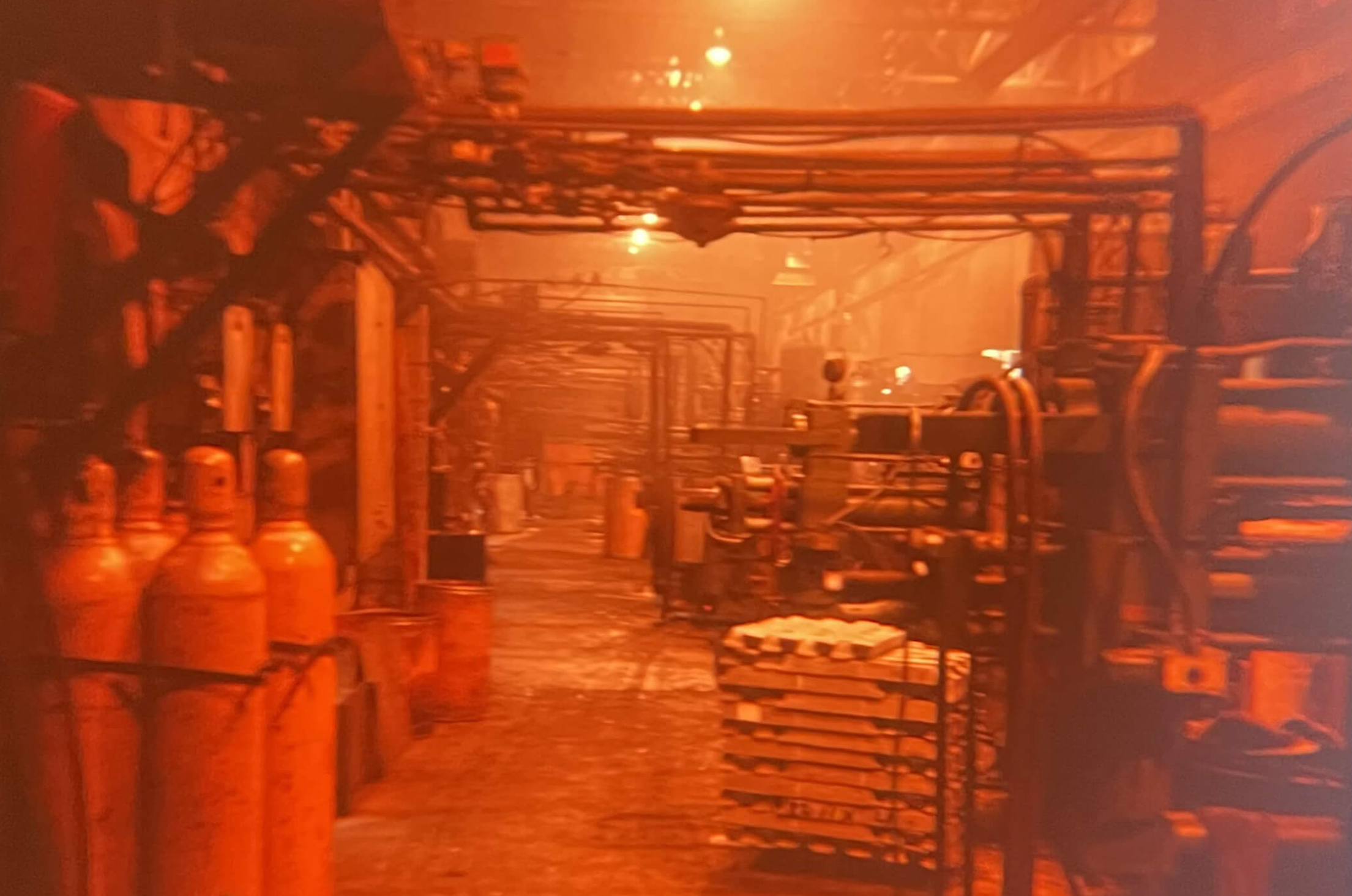 Carpenter Die Casting Rows of industrial gas cylinders, machinery, and metal stacks line a dimly lit factory floor with orange lighting.