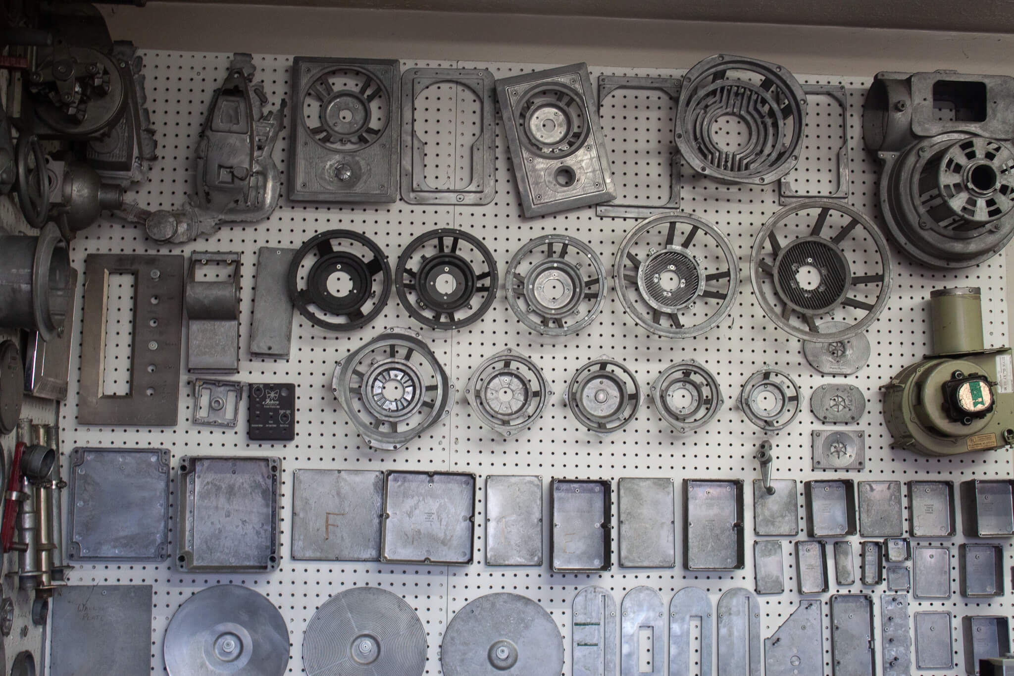 Carpenter Die Casting Assorted metal machine parts and components are arranged neatly on a white pegboard wall in a workshop or garage setting.