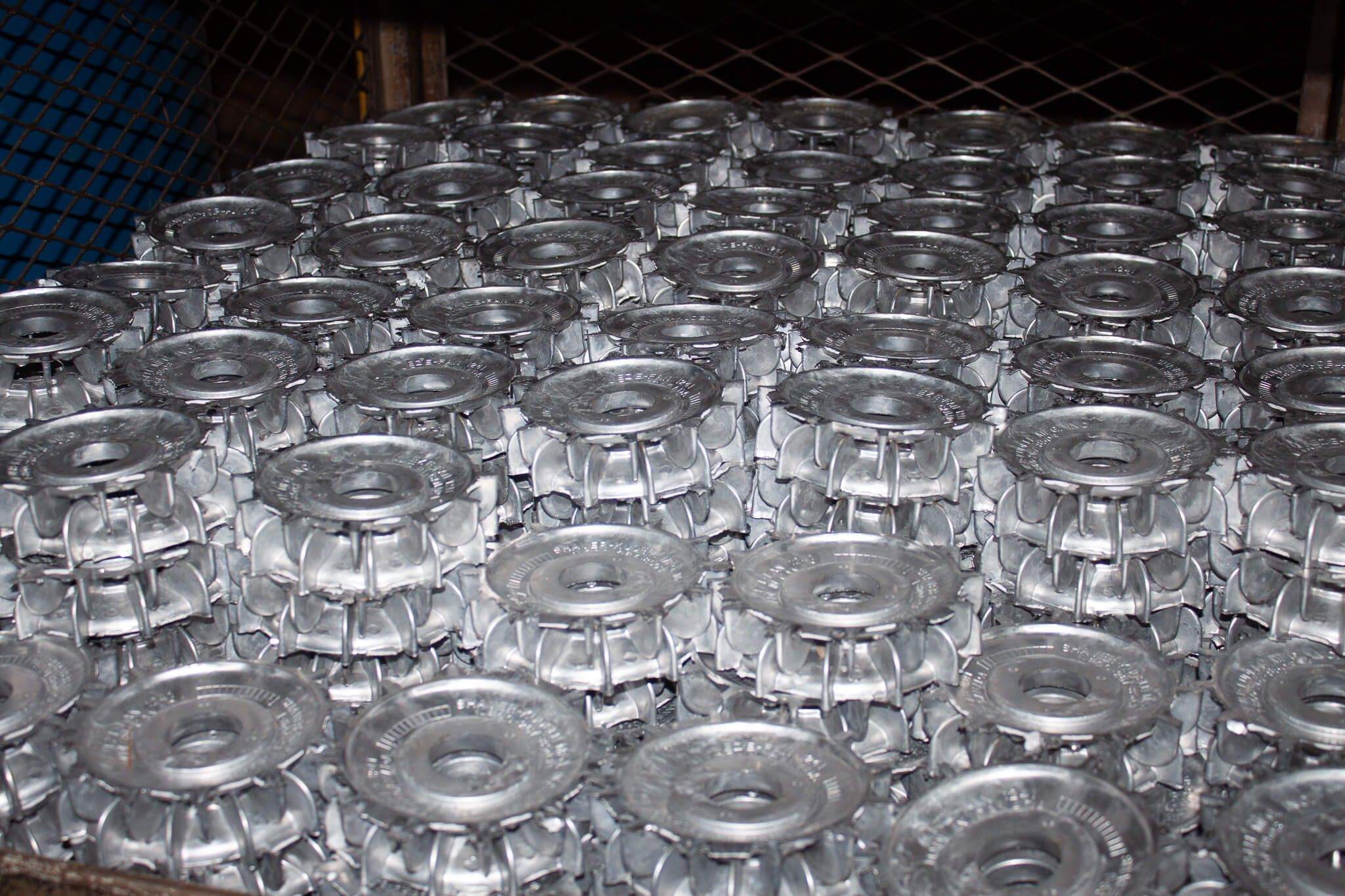 Carpenter Die Casting Rows of identical silver metal machine parts are stacked in a grid on a metal surface, with a wire mesh visible in the background.