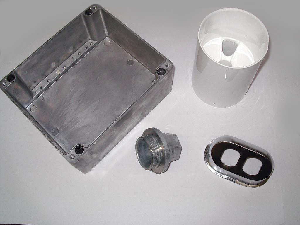 Carpenter Die Casting Four separate industrial parts including a metal box, a white cylindrical housing, a threaded metal connector, and a plastic or metal faceplate are arranged on a white surface.